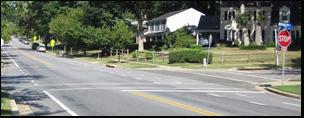 Photo shows a 2-lane street with a shoulder on the left and bike lane and parking lane on the right.  Nearest to us is an intersection where our street has a stop sign.  About 200 feet ahead is a yellow sign and 150 feet ahead of that is another yellow sign -- we cannot read what the yellow signs say.